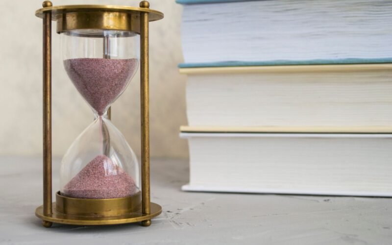 Close-up of hourglass with red sand beside stacked books, symbolizing time and knowledge.