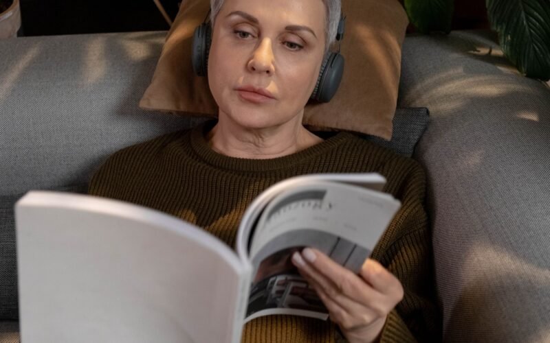 A woman in headphones reading a magazine on a couch in a cozy indoor setting.