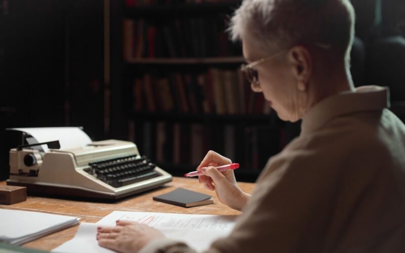 Elderly woman reviewing a manuscript with a typewriter at a wooden desk.