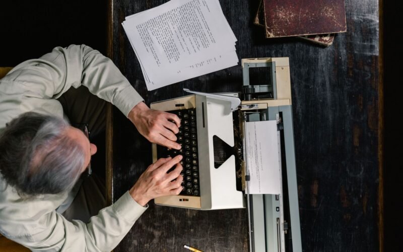 A person typing on a vintage typewriter with scattered papers on a wooden table.